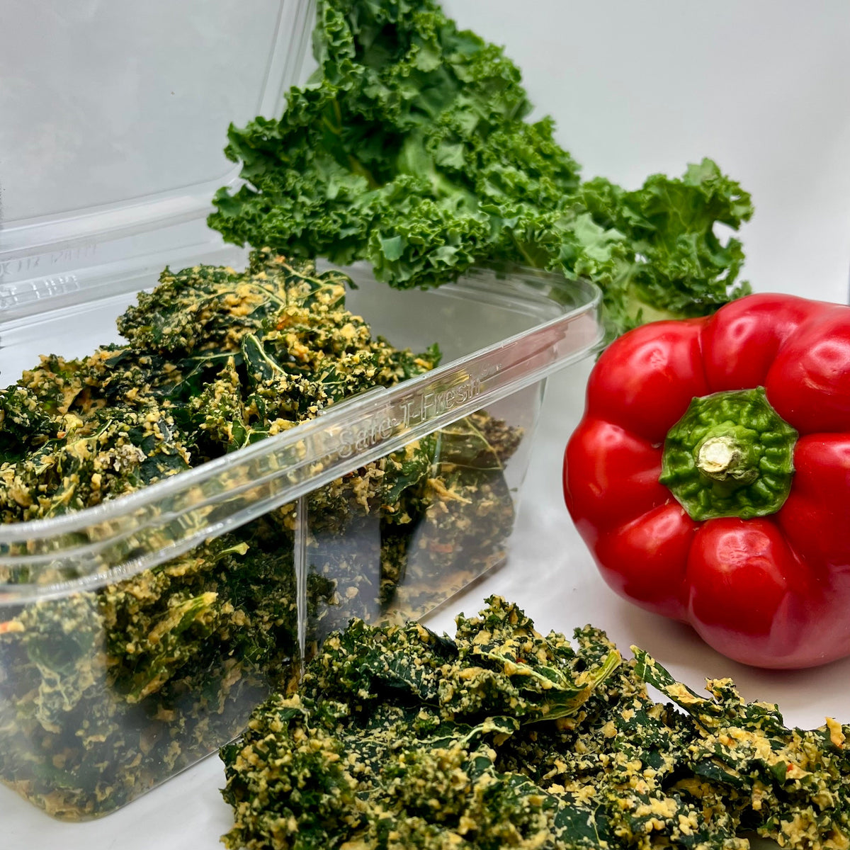 Kale chips in a clear container with a red bell pepper on a white background