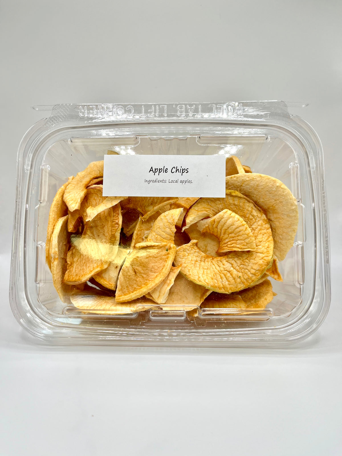 A clear plastic container of Healing Home Foods Apple Chips sits against a white background. The lid’s label reads: Apple Chips and Ingredients: Local apples, highlighting these delicious dehydrated apples.