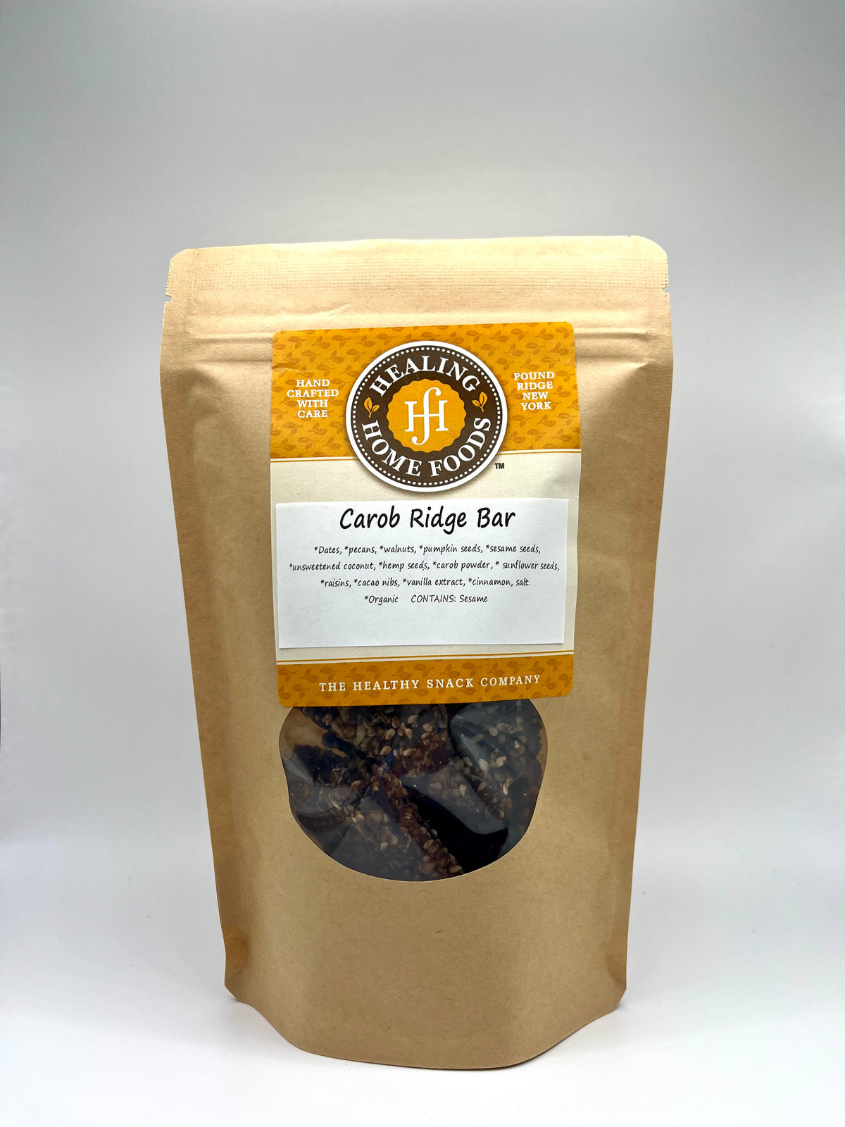 A brown resealable pouch labeled Healing Home Foods Carob Ridge Bar features a round window showing the snack bar inside. The packaging notes it is handcrafted and made with organic ingredients, including carob, healthy nuts, and seeds.