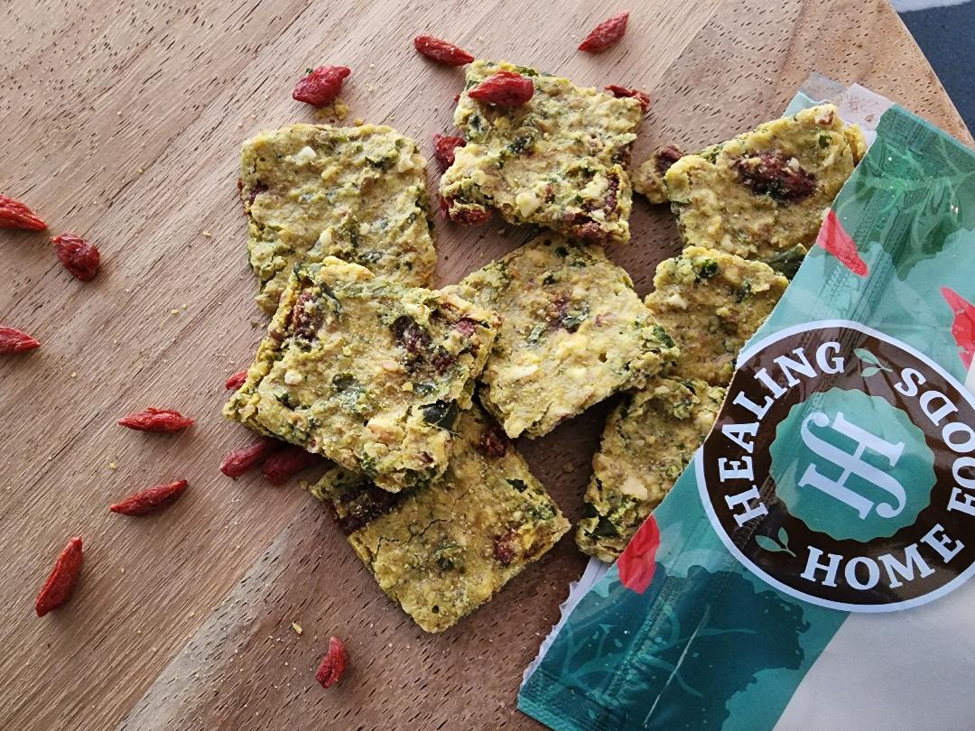 Rectangular yellow-green Healing Home Foods Kale Goji Crackers with red goji berries are spread on a wooden surface next to an open teal and white package. Loose goji berries surround these air-dried superfood crackers. Case of 8.